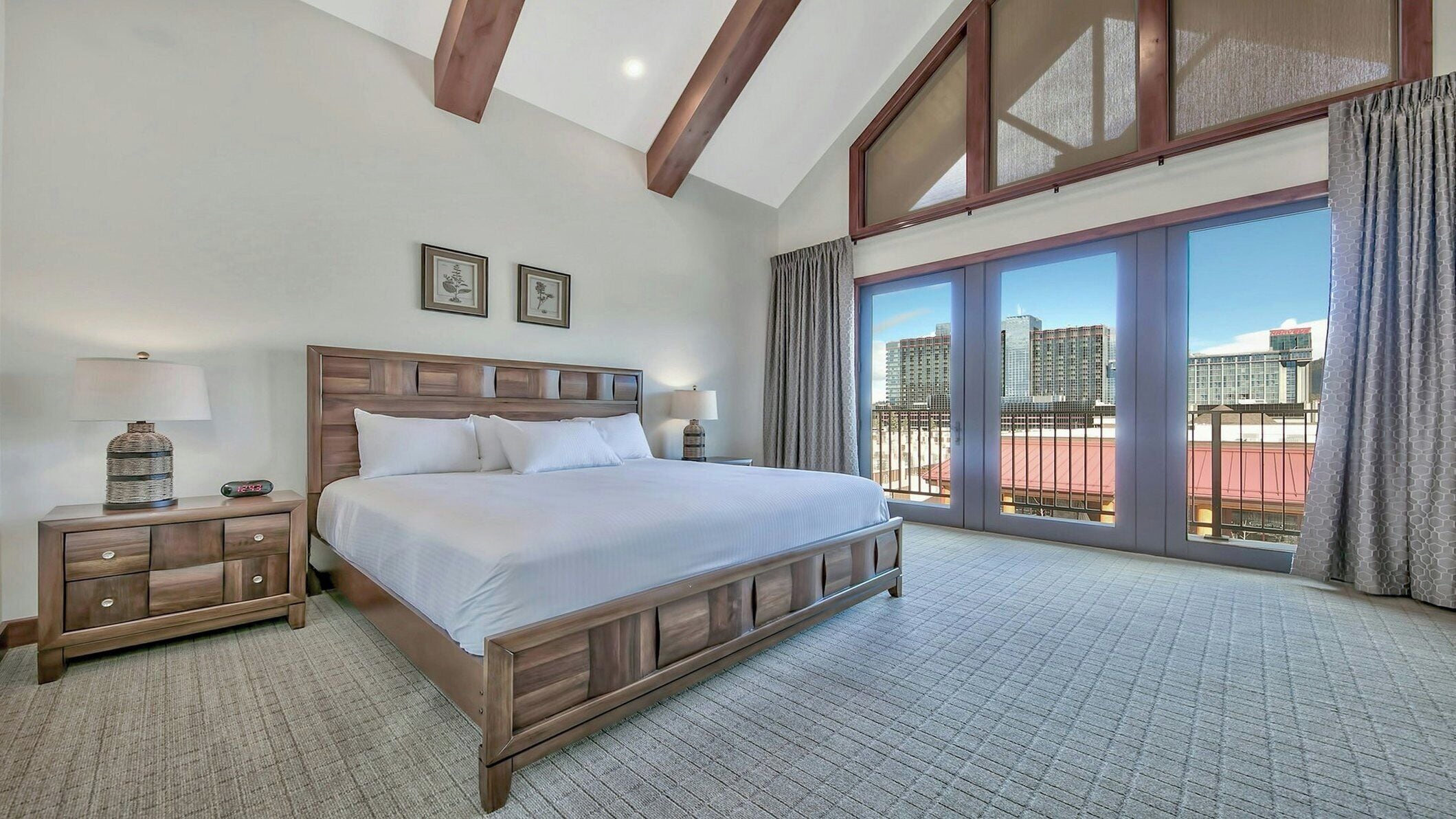 A bedroom with a large bed, two bedside tables, a nightstand, and a view of the city through large windows.