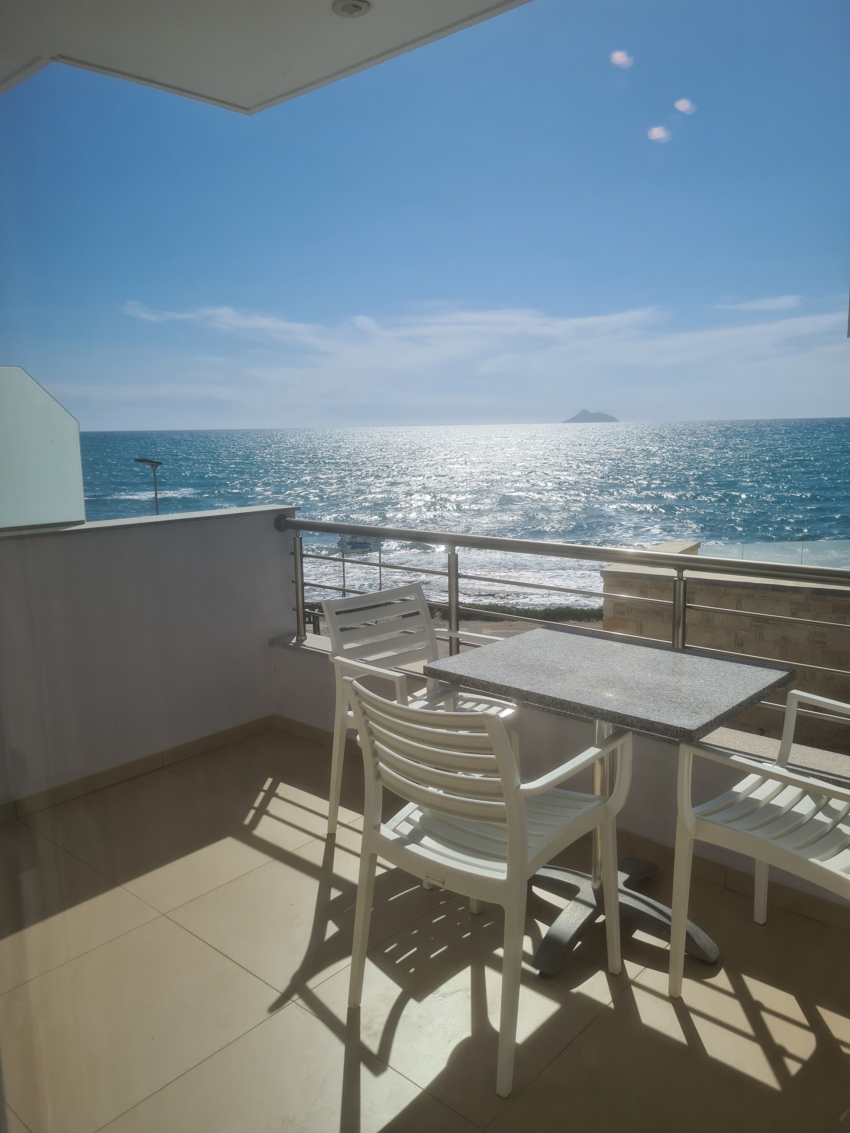 Un balcone esterno con tavolo e sedie, affacciato su un mare azzurro e brillante con un'isola in lontananza.