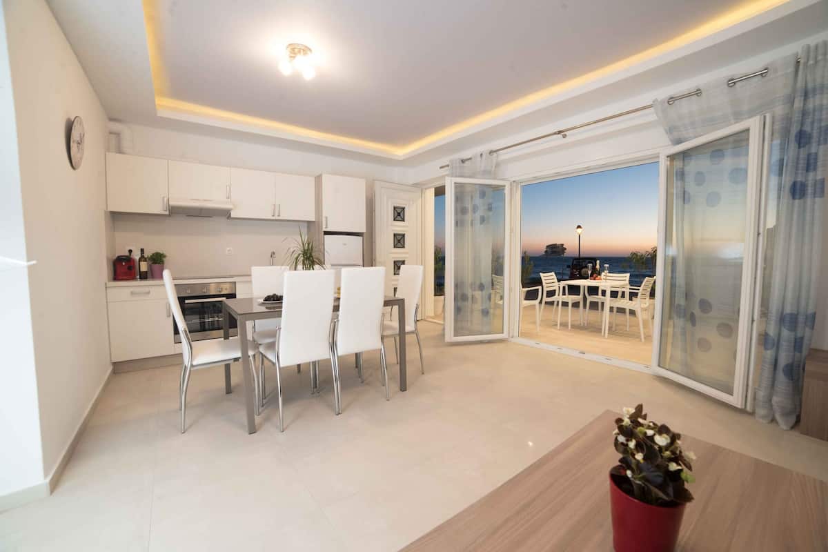Een moderne keuken met witte kasten, een eethoek met witte stoelen en uitzicht op zee door grote glazen deuren.