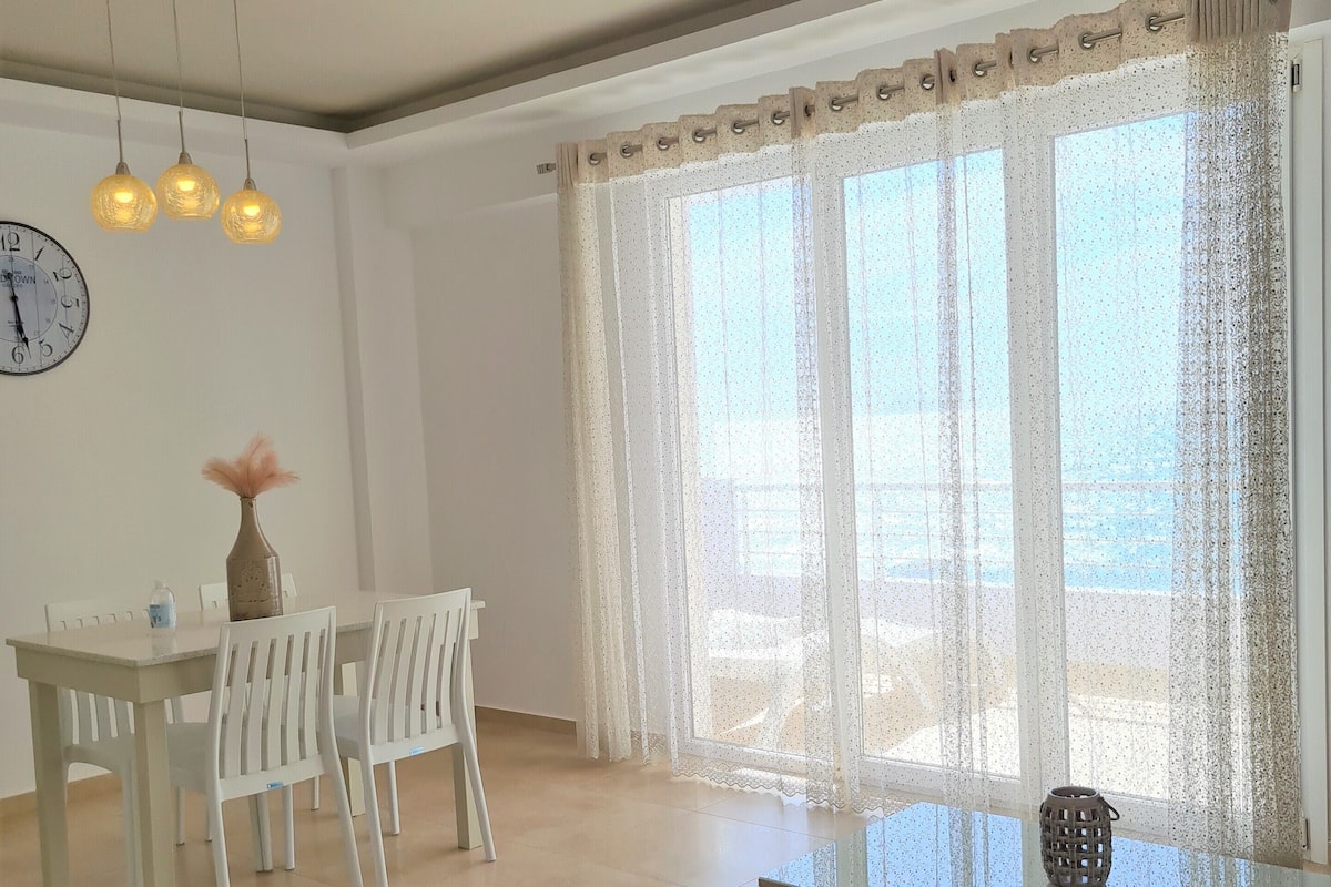 Een moderne woonkamer met een bank, eettafel, stoelen en een groot raam met dunne gordijnen.