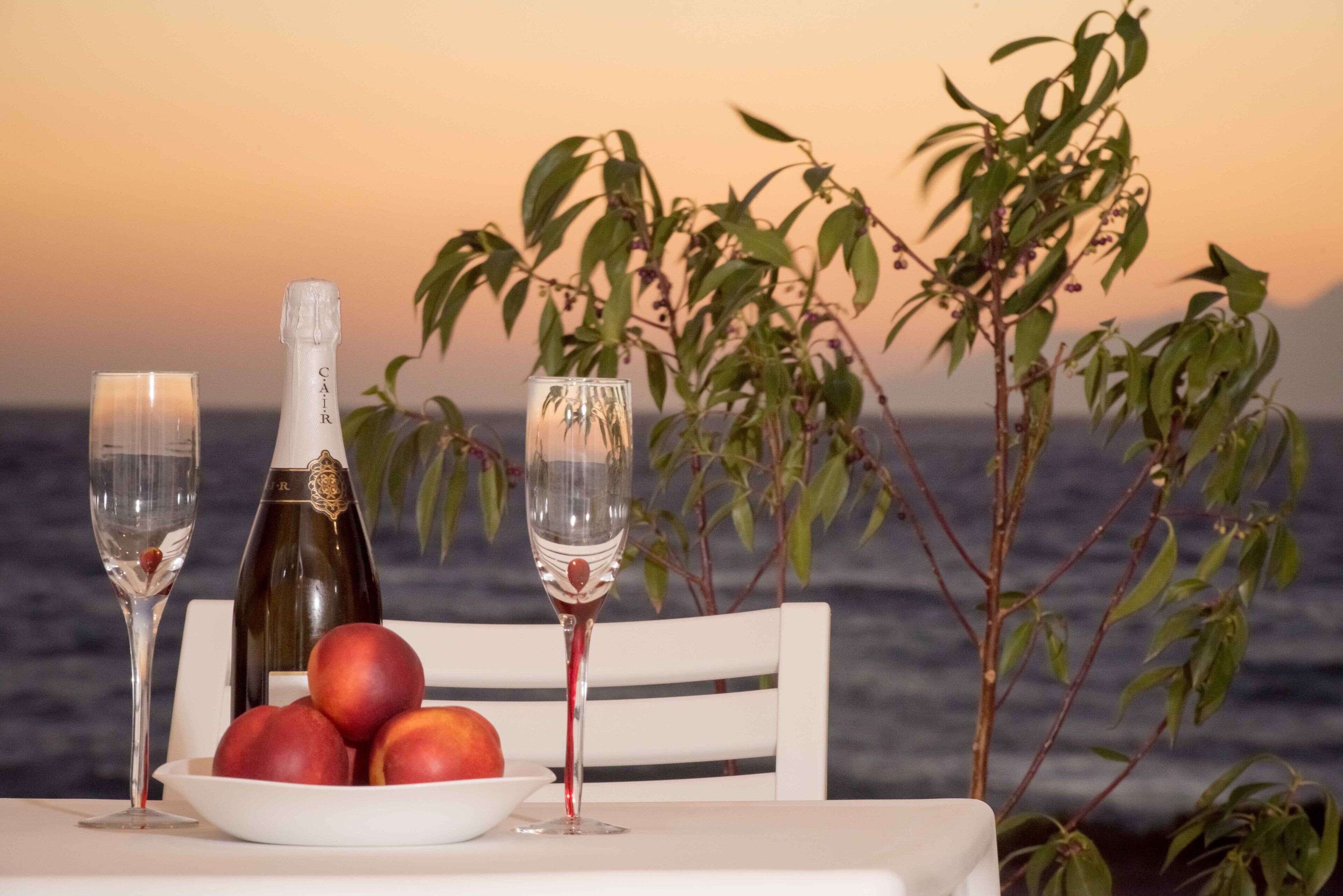 A table with a bottle of champagne, two glasses, and a bowl of fruit by a window overlooking the sea.