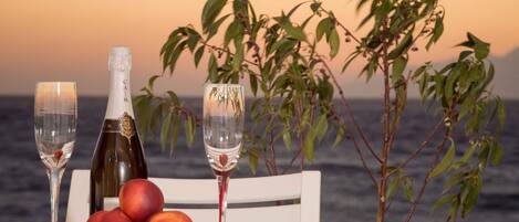 A table with a bottle of champagne, two glasses, and a bowl of fruit by a window overlooking the sea.