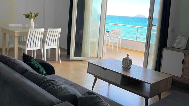 A modern living room with a sofa, a dining table with chairs, and a view of the sea through an open door.