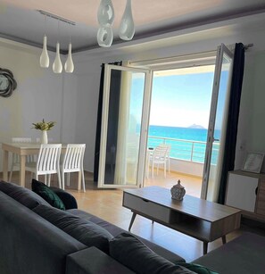 A modern living room with a sofa, a dining table with chairs, and a view of the sea through an open door. - Bella Mare Luxury Apartments (Faistos)