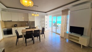 A modern kitchen with white cabinets, a dining area with a table and chairs, a TV on a stand, and a large sliding glass door leading to a balcony with a view of the sea.