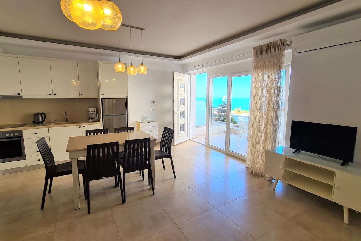 Een moderne keuken met witte kastjes, een eethoek met tafel en stoelen, een televisie op een standaard en een grote schuifpui die leidt naar een balkon met uitzicht op zee.