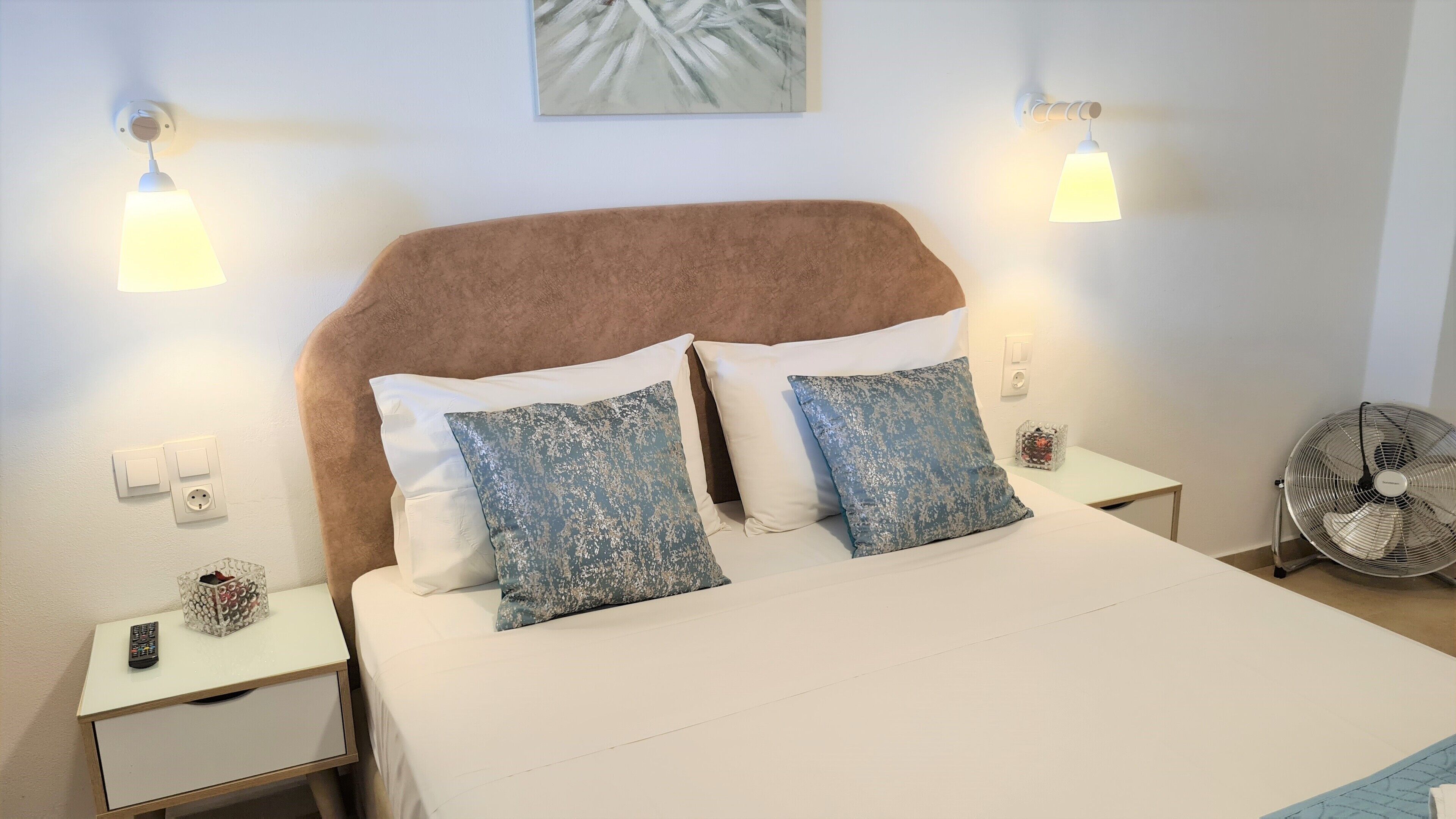 Una cama bien hecha con un cabecero, dos mesitas de noche con lámparas, un ventilador en la pared y un cuadro sobre la cama.