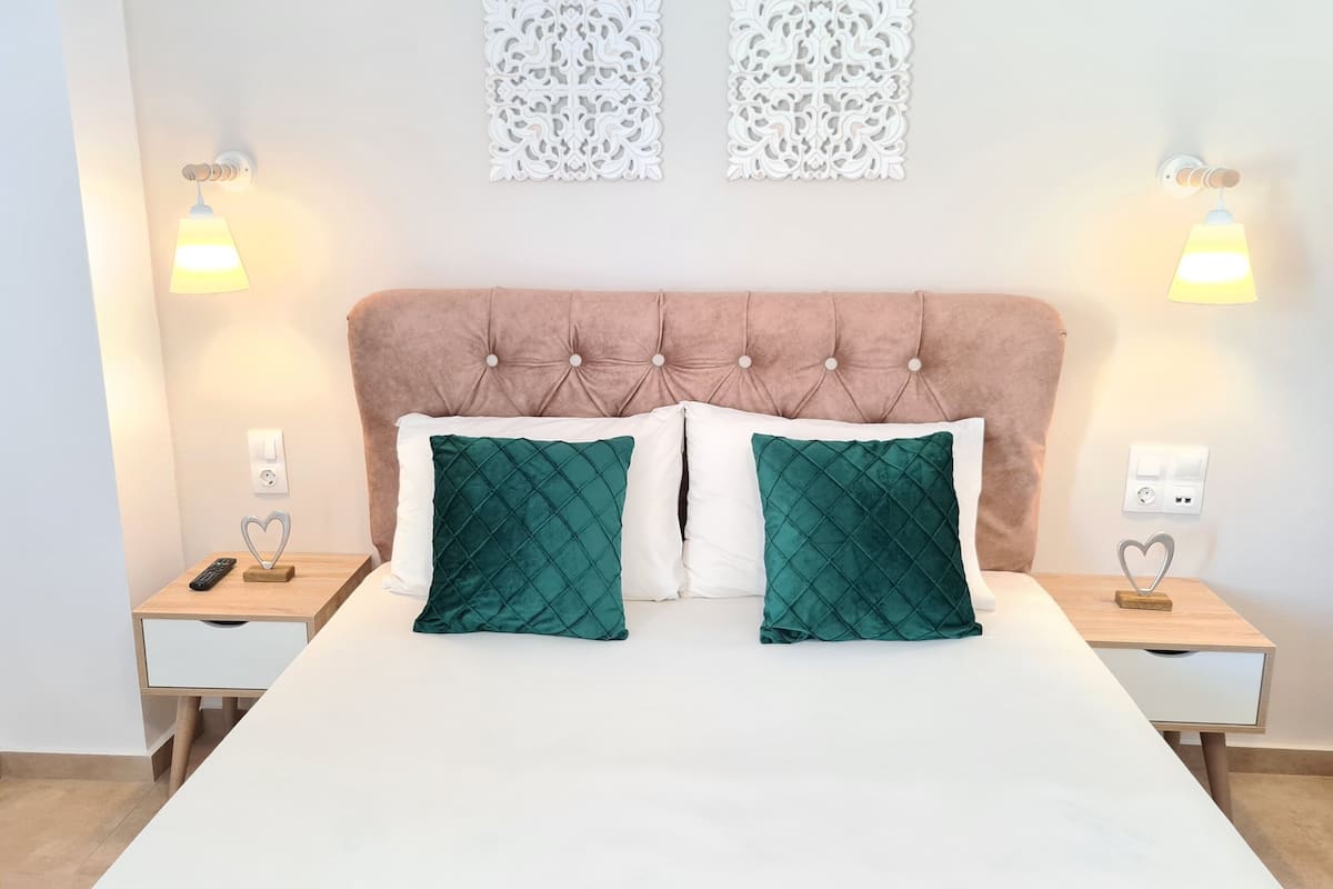 Een slaapkamer met een gestoffeerd hoofdeinde, twee nachtkastjes, een bed met witte en groene kussens en decoratieve wandkunst.