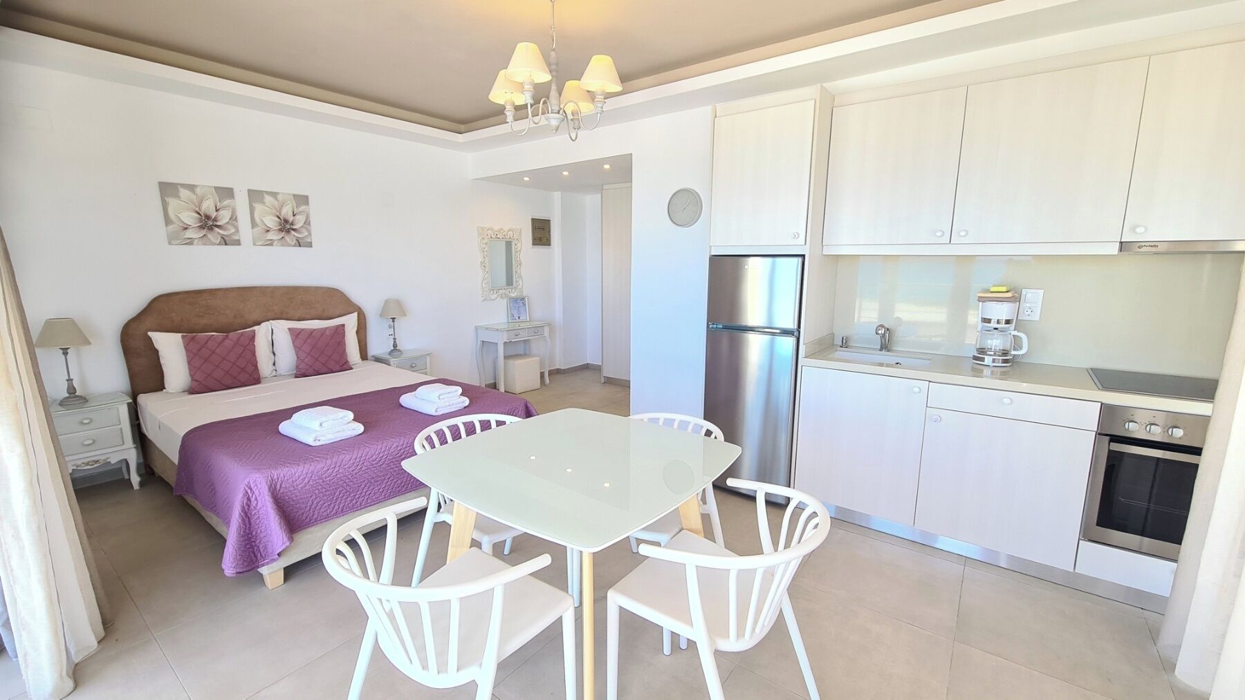 A bedroom with a bed, bedside tables, and a nightstand. There is a dining area with a table and chairs. The kitchen has cabinets, a refrigerator, and a stove.