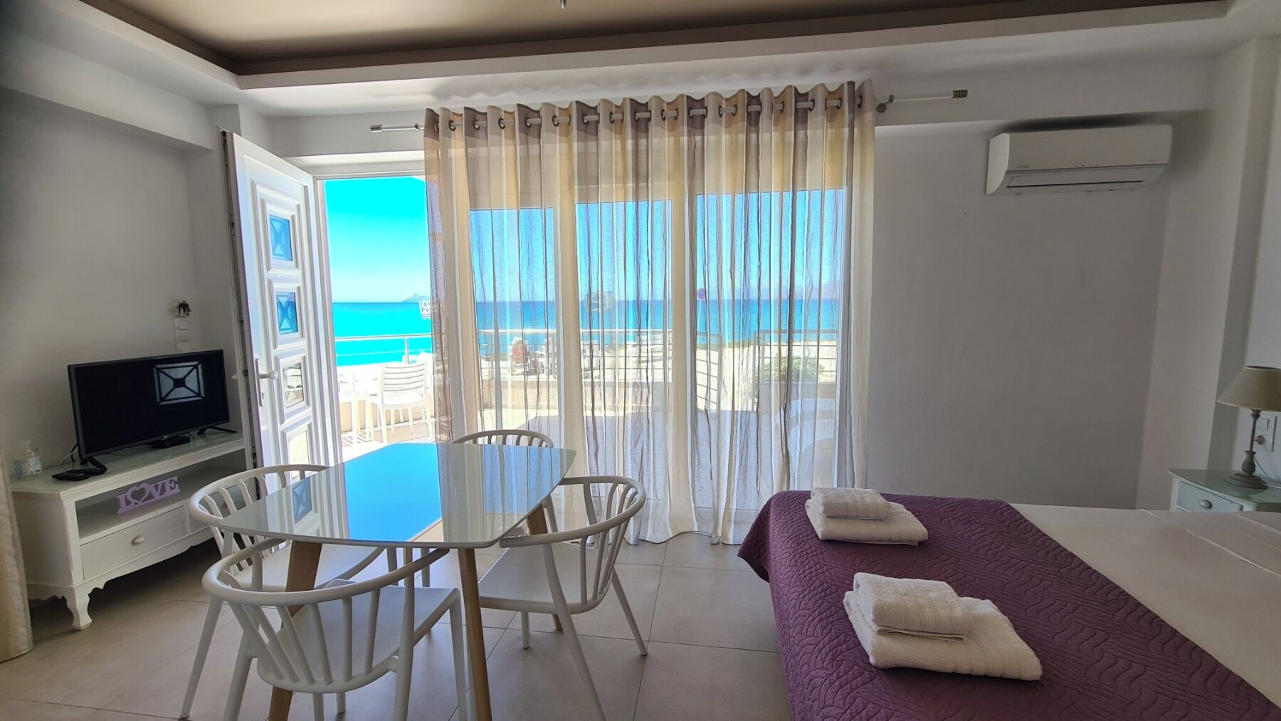 Une chambre d’hôtel avec un balcon, un lit recouvert d’une couverture violette, un téléviseur posé sur un meuble blanc et une table avec des chaises.