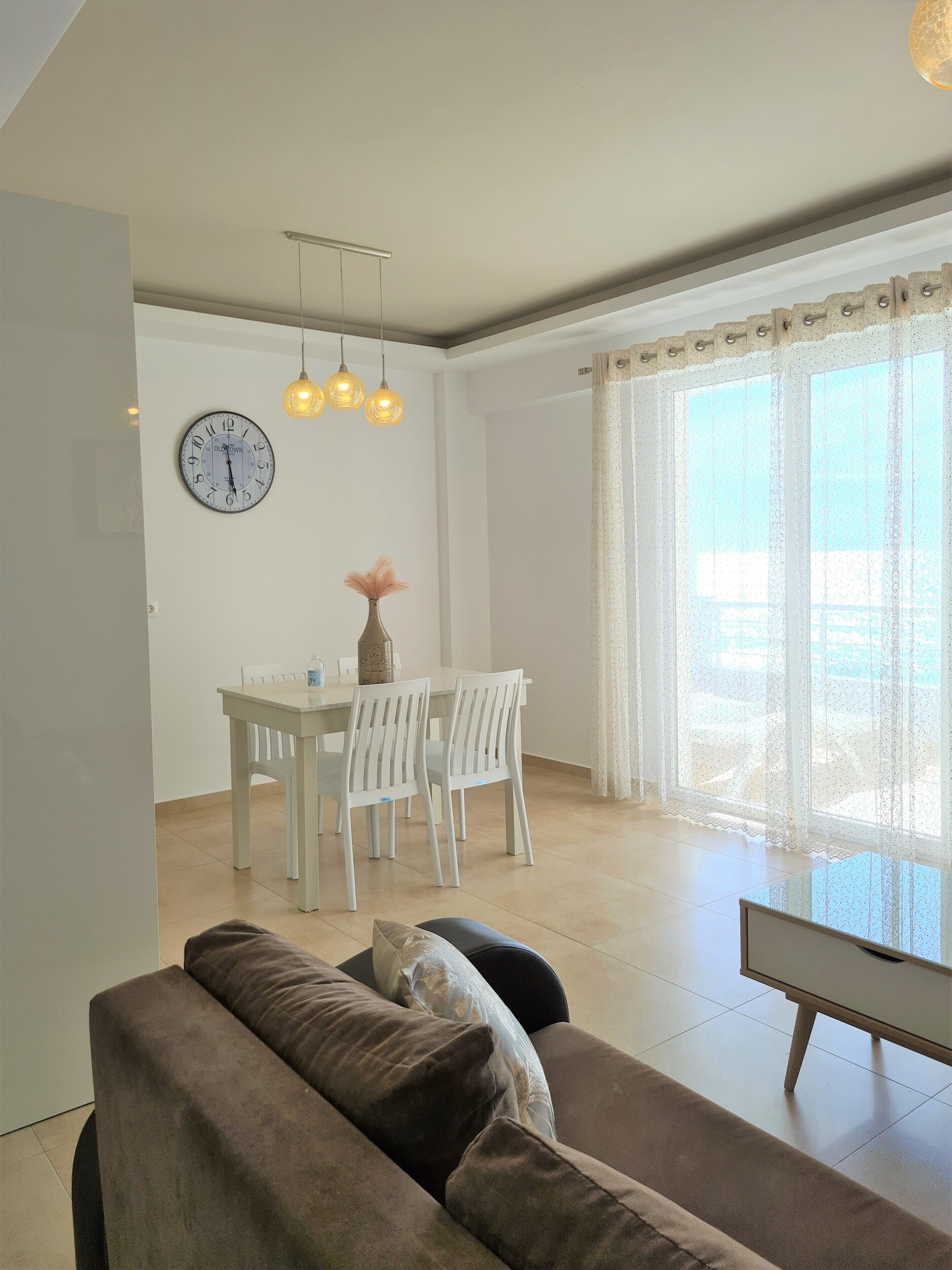 Ein modernes Wohnzimmer mit einem braunen Sofa, einem Esstisch mit Stühlen, einer Wanduhr und einem großen Fenster mit transparenten Vorhängen.