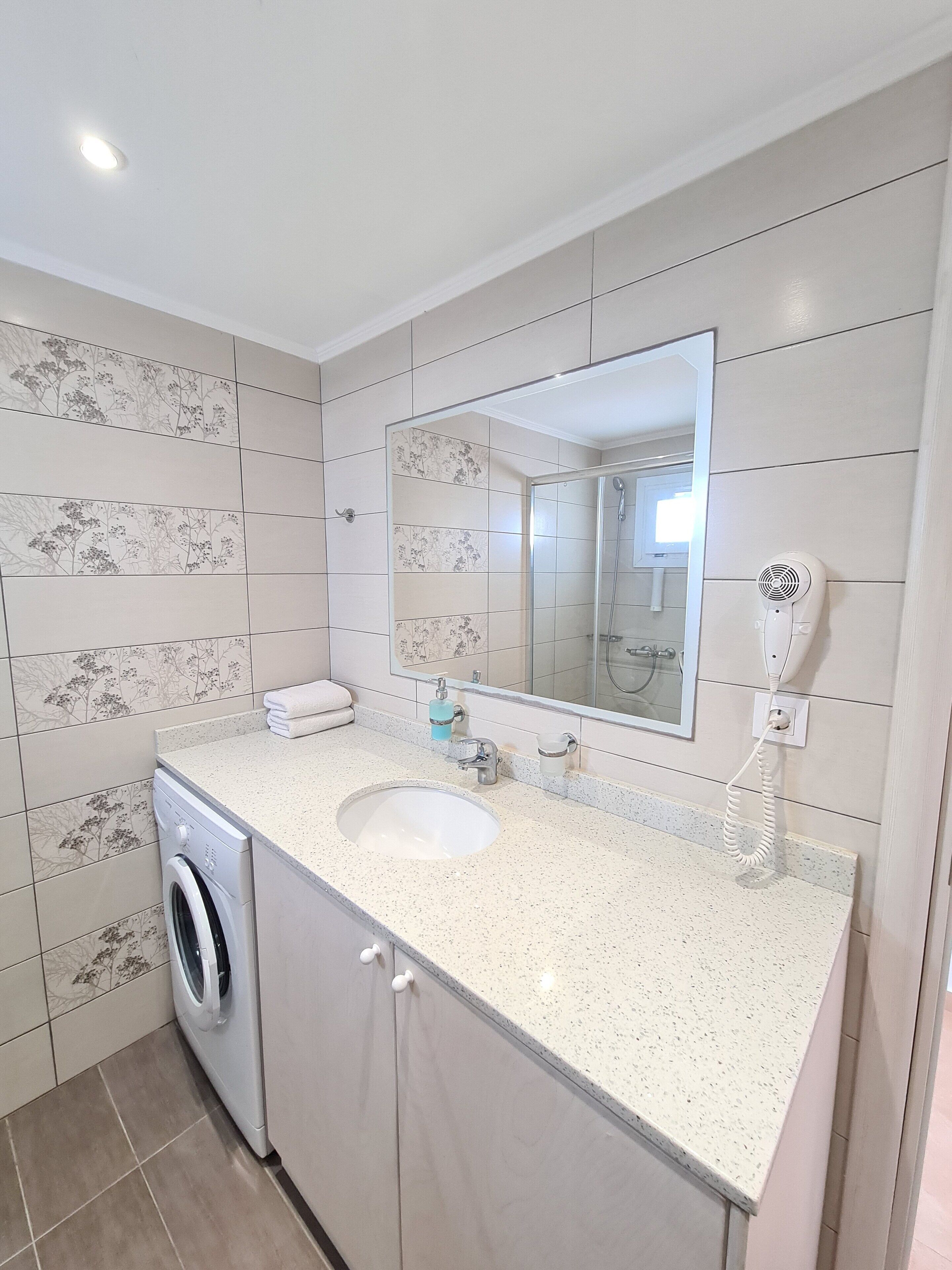 A bathroom with a washing machine, a sink, a mirror, and a hairdryer.