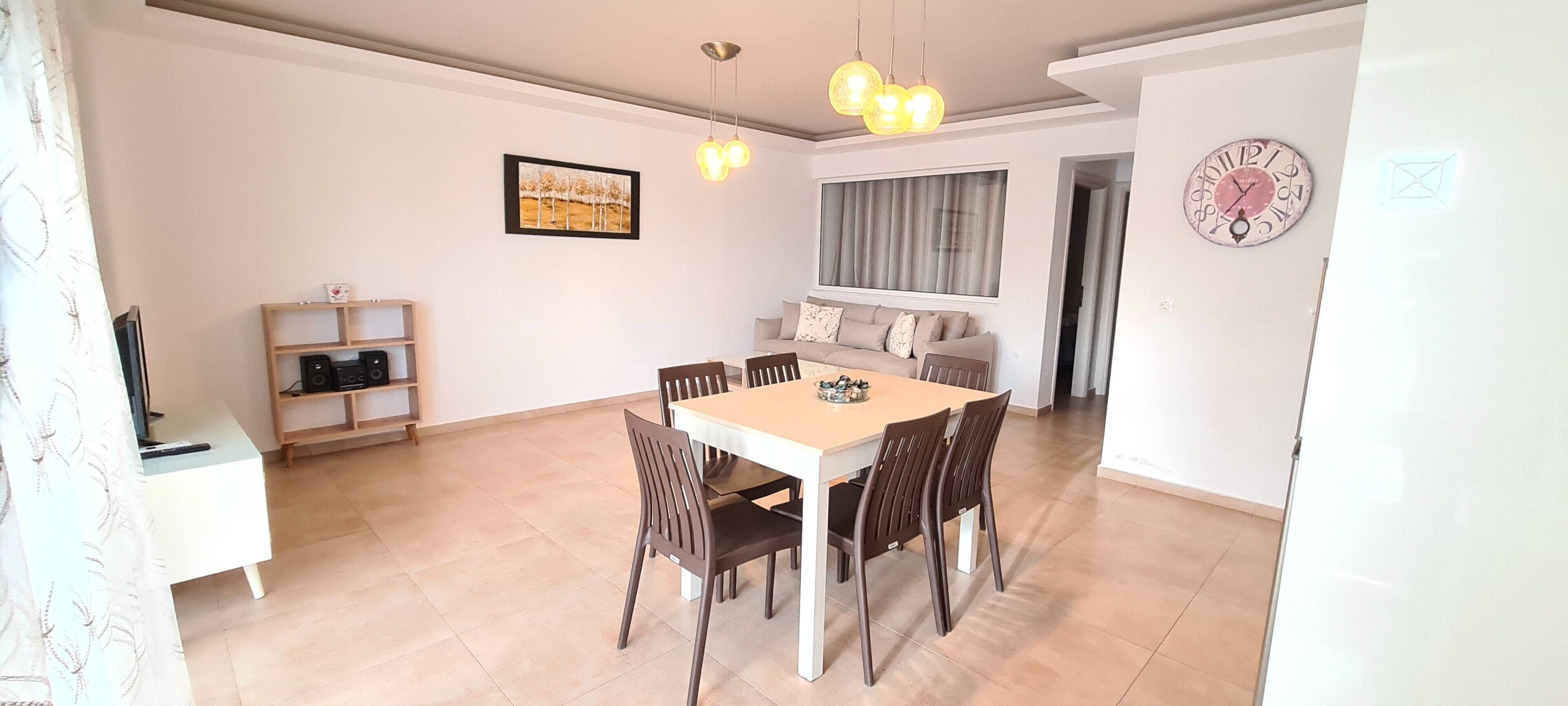 A dining area with a white table and brown chairs, a bookshelf, a painting on the wall, and a clock.