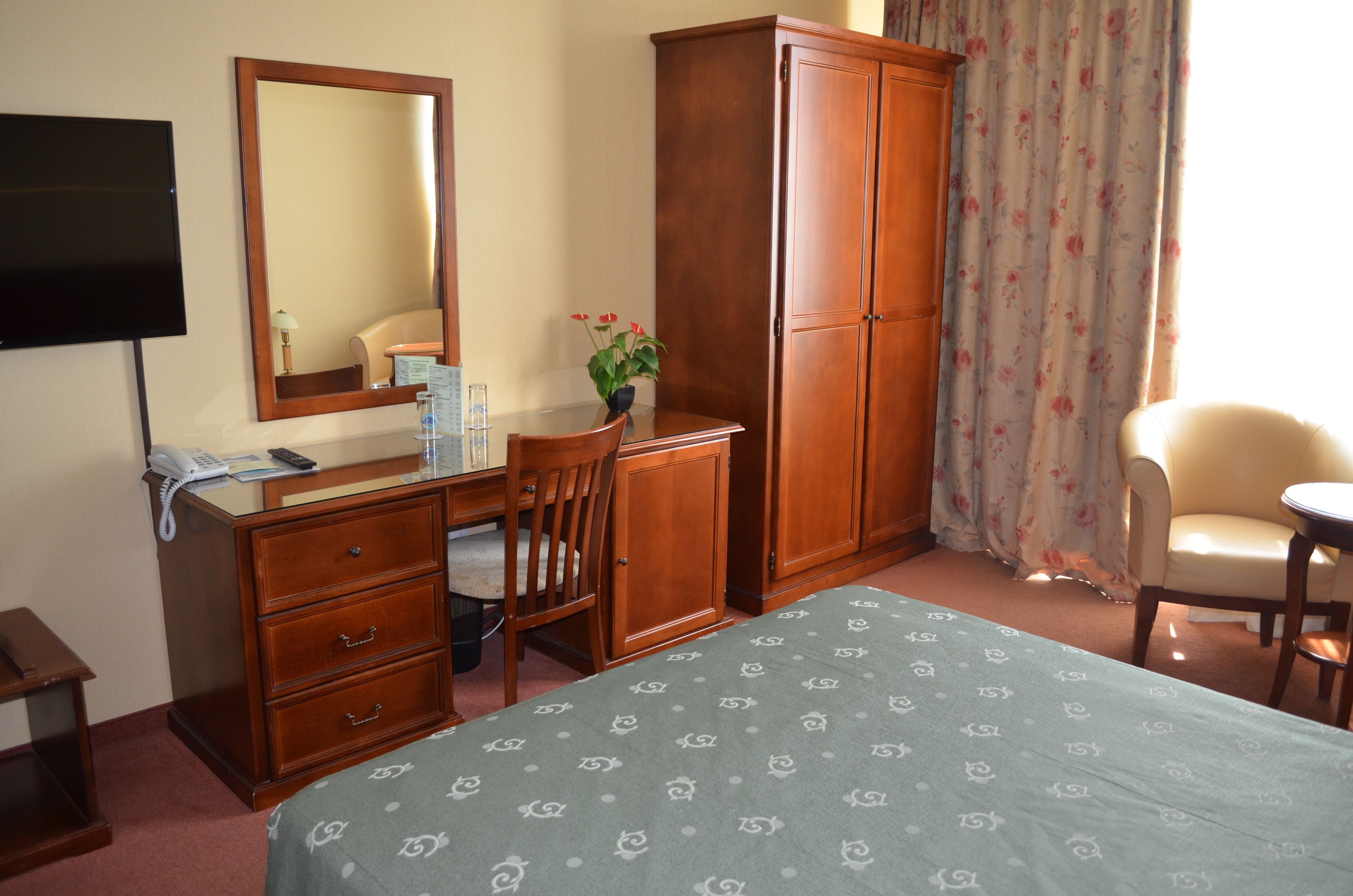 A hotel room with a bed, a desk with a chair, a wardrobe, a television, and a window with curtains.