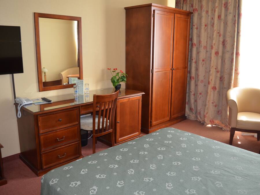 A hotel room with a bed, a desk with a chair, a wardrobe, a television, and a window with curtains.