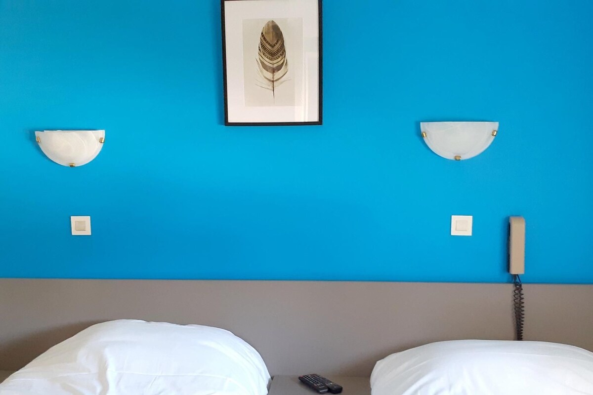 Une chambre d’hôtel avec un mur bleu, deux tables de chevet, un lit avec une literie blanche, une œuvre d’art représentant des plumes encadrée, un téléphone et une télécommande sur la table de chevet.