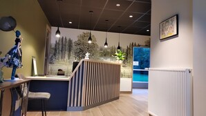 Uma área de recepção com balcão de madeira, espaço de estar com uma poltrona, iluminação pendente, um mural na parede e um radiador.