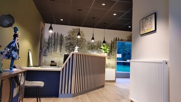 Zona de recepción con mostrador de madera, área de estar con una silla, iluminación colgante, un mural en la pared y un radiador.