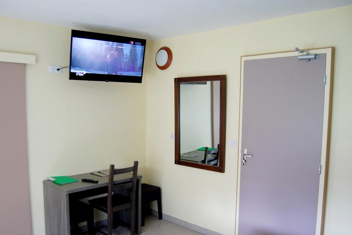 Una habitación con una televisión montada en la pared, un escritorio con una silla, un espejo y una puerta.