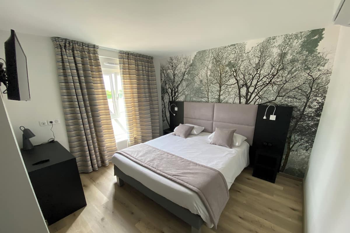Een moderne slaapkamer met een groot bed, een nachtkastje, een aan de muur bevestigde televisie en een muurschildering van bomen.
