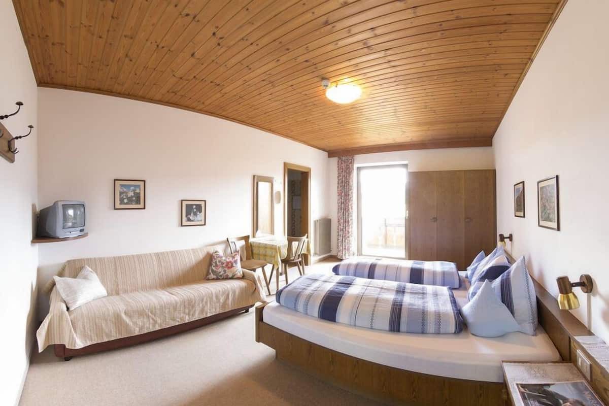 A cozy bedroom with a wooden ceiling, a bed with blue and white bedding, a sofa, a television, and framed pictures on the walls.