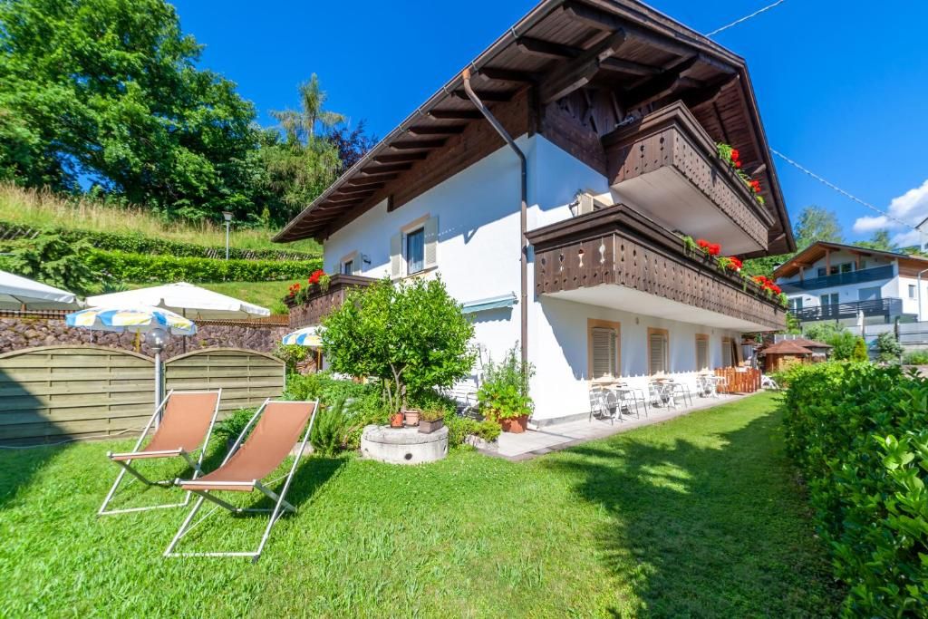 A two-story chalet with a sloped roof, a balcony with red flowers, and a well-maintained garden with lawn chairs.
