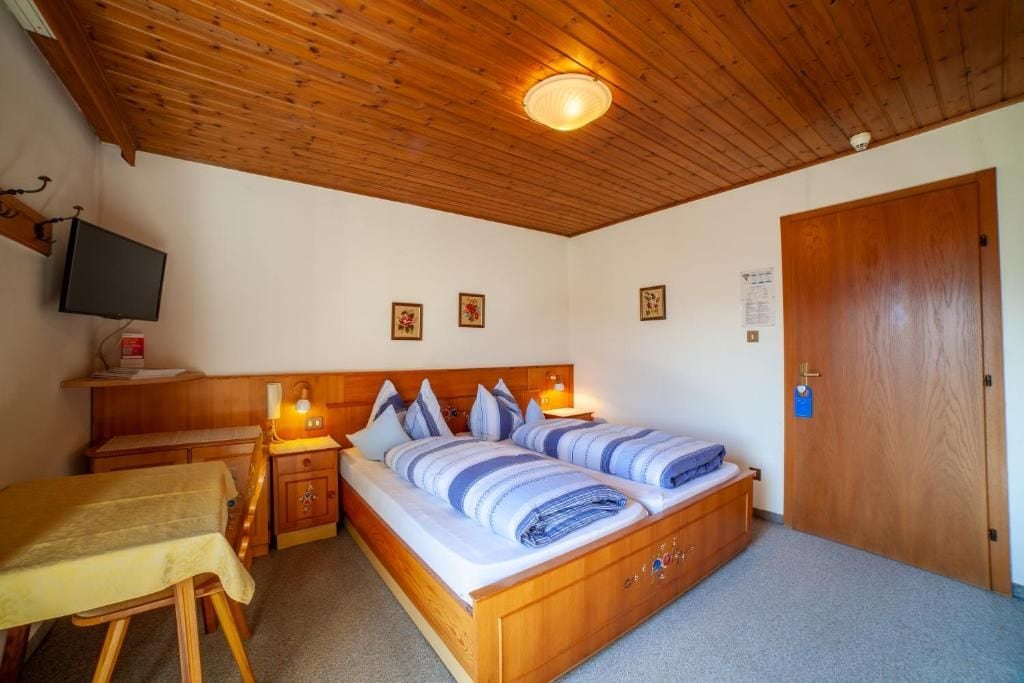 A bedroom with a wooden ceiling, a bed with blue bedding, a bedside table with a lamp, and a television on the wall.