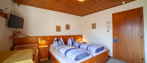 A bedroom with a wooden ceiling, a bed with blue bedding, a bedside table with a lamp, and a television on the wall.