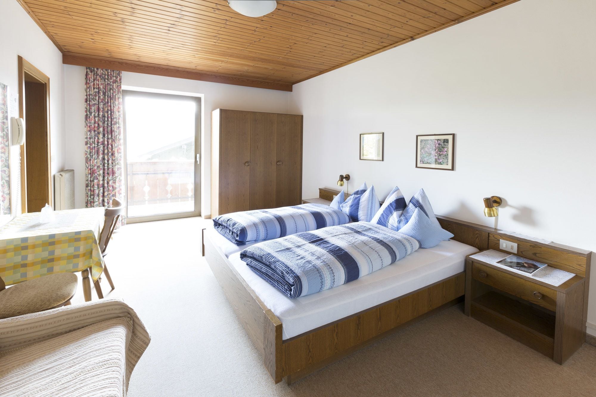 Ein Schlafzimmer mit Holzboden, einem großen Bett mit blau-weißer Bettwäsche, einem Holzschrank, einem Nachttisch mit Lampe und einem Fenster mit Vorhängen.