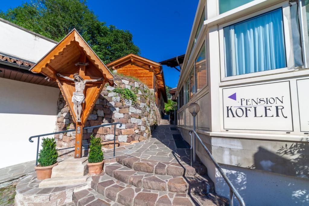 Un edificio con una escalera de piedra que conduce a una estructura de madera con una cruz. Hay un letrero que dice 'Pension Kofler' en el edificio.
