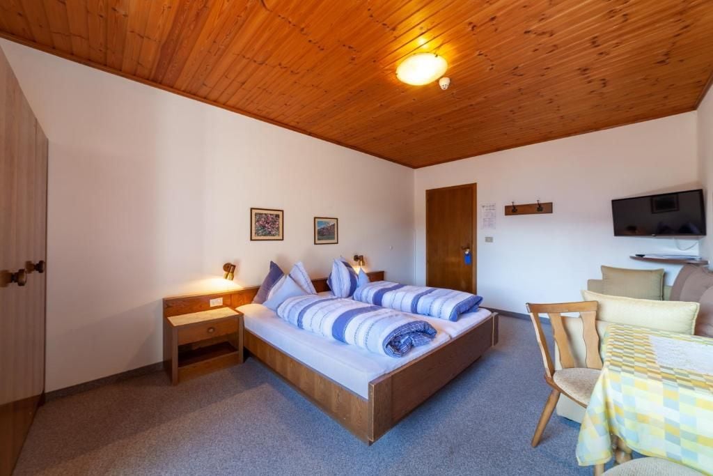 Une chambre avec un plafond en bois, un lit avec une literie à rayures, une table de chevet, une chaise, une petite table recouverte d’une nappe à carreaux et un téléviseur fixé au mur.