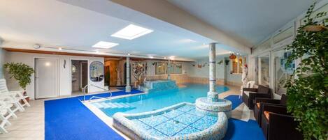 An indoor swimming pool with a blue mosaic border, a water fountain, and seating areas with plants.