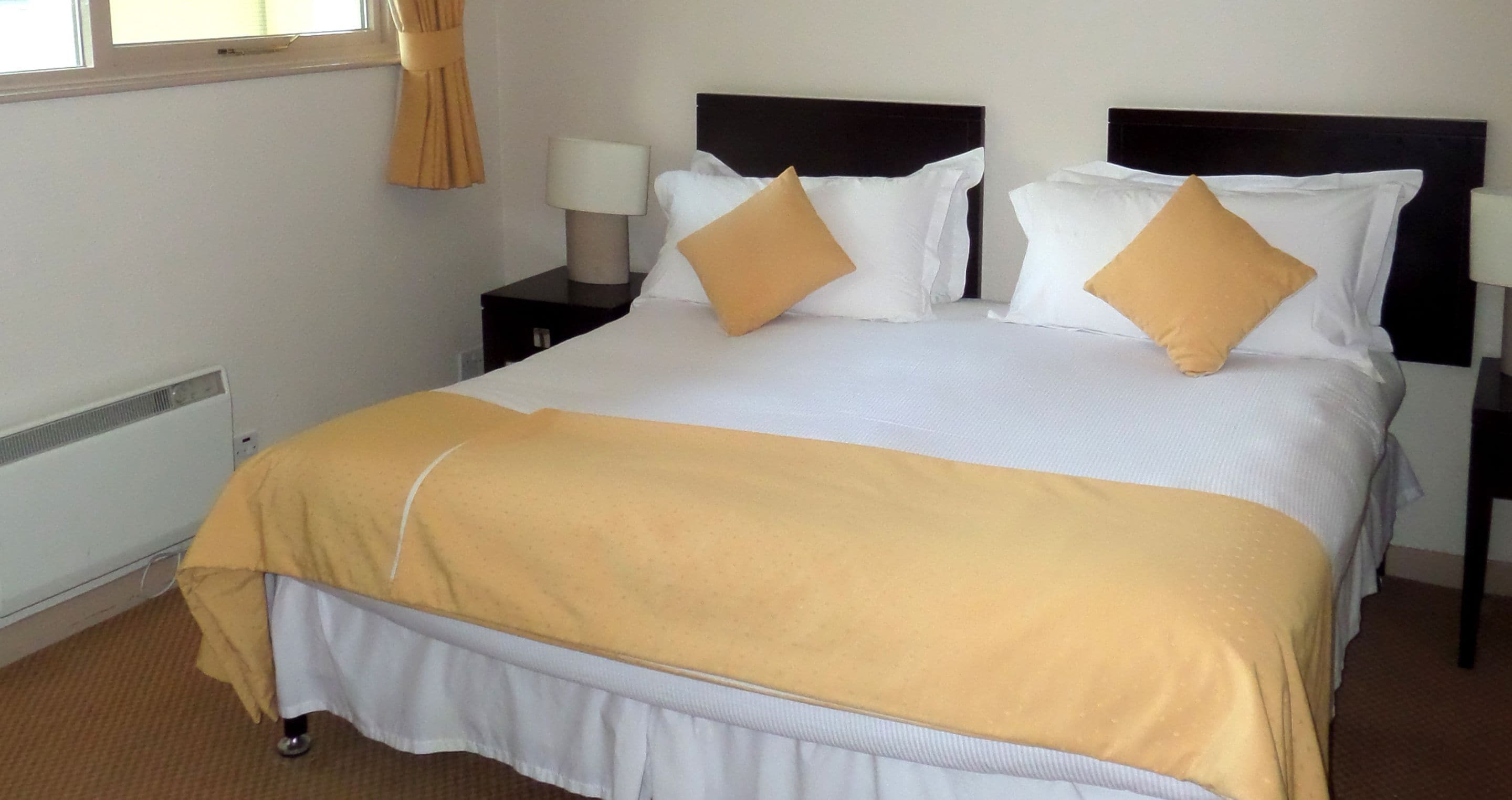 A neatly made bed with a white and yellow bedspread, two bedside tables with lamps, and a window with curtains.