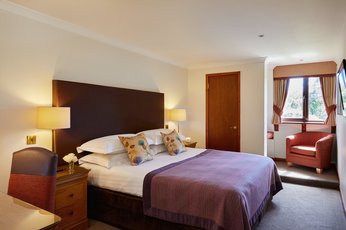 A hotel room with a bed, bedside lamps, a chair, a desk, and a window with curtains.