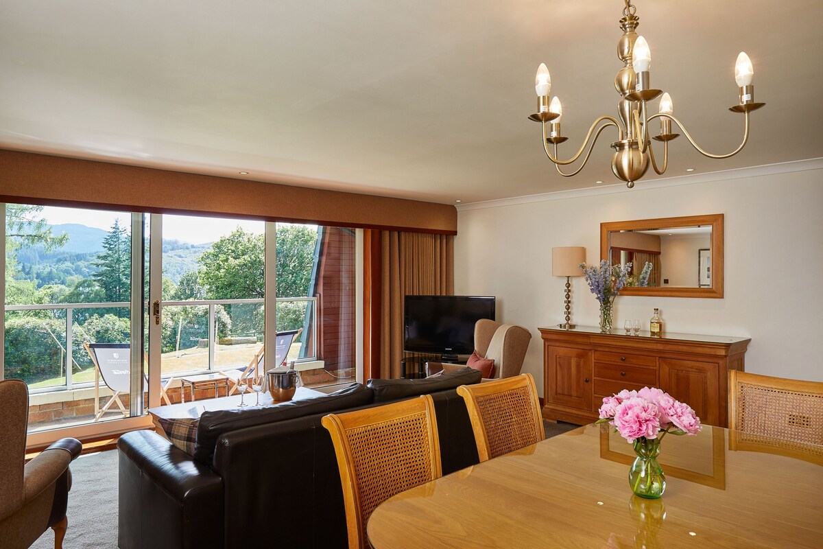 A spacious living area with a large window, a sofa set, a dining table, and a television.