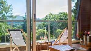 Una habitación con ventanas amplias, dos sillones, una mesita con una copa de vino y un escritorio de madera con un periódico.