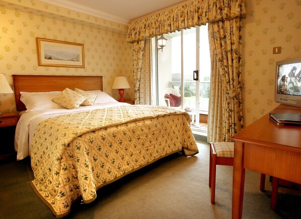 Habitación de hotel con cama, escritorio con televisor, ventana con cortinas, mesita de noche con lámpara y papel tapiz floral en la pared.