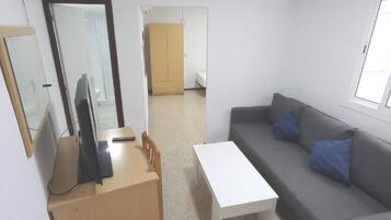 A room with a grey sofa, a white table, a wooden desk, a mirror, and a wardrobe.