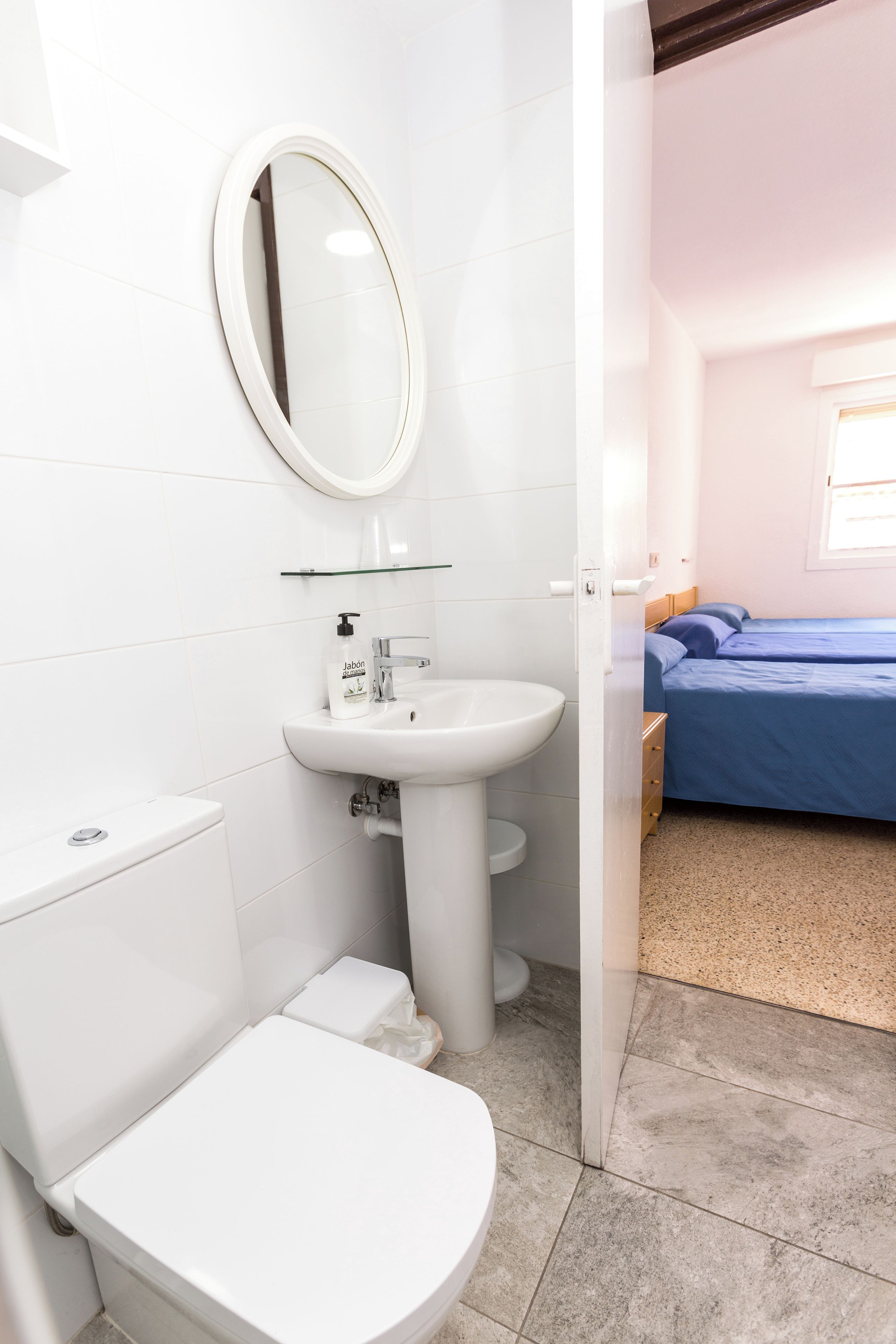 A bathroom with a toilet, sink, and mirror. There is a bed with blue bedding in the adjacent room.