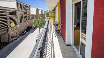 Un balcon donnant sur des vélos et des scooters garés, un petit arbre et une table avec des chaises.
