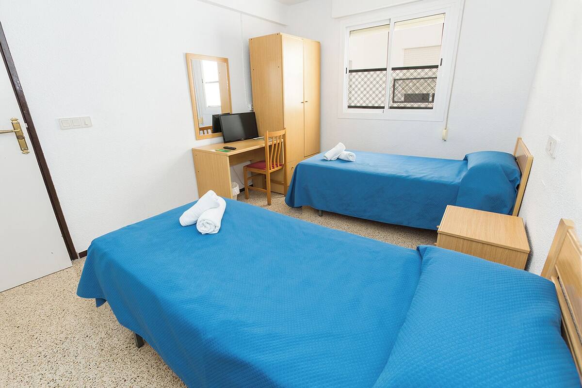 Habitación de hotel con dos camas individuales, un escritorio con silla, una computadora y ventana con vista al exterior.