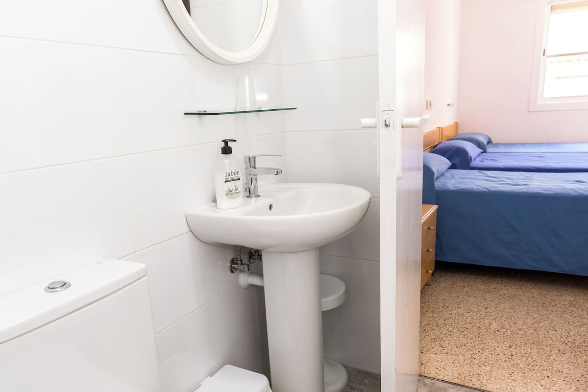 Baño con inodoro, lavabo y espejo. En la habitación contigua, hay una cama con ropa de cama azul.