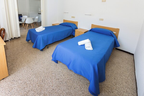 Une chambre d’hôtel avec deux lits simples, chacun recouvert d’une couverture bleue et agrémenté d’une serviette blanche. Une table de chevet en bois se trouve entre les lits, et une petite table avec des chaises est visible au fond de la pièce.