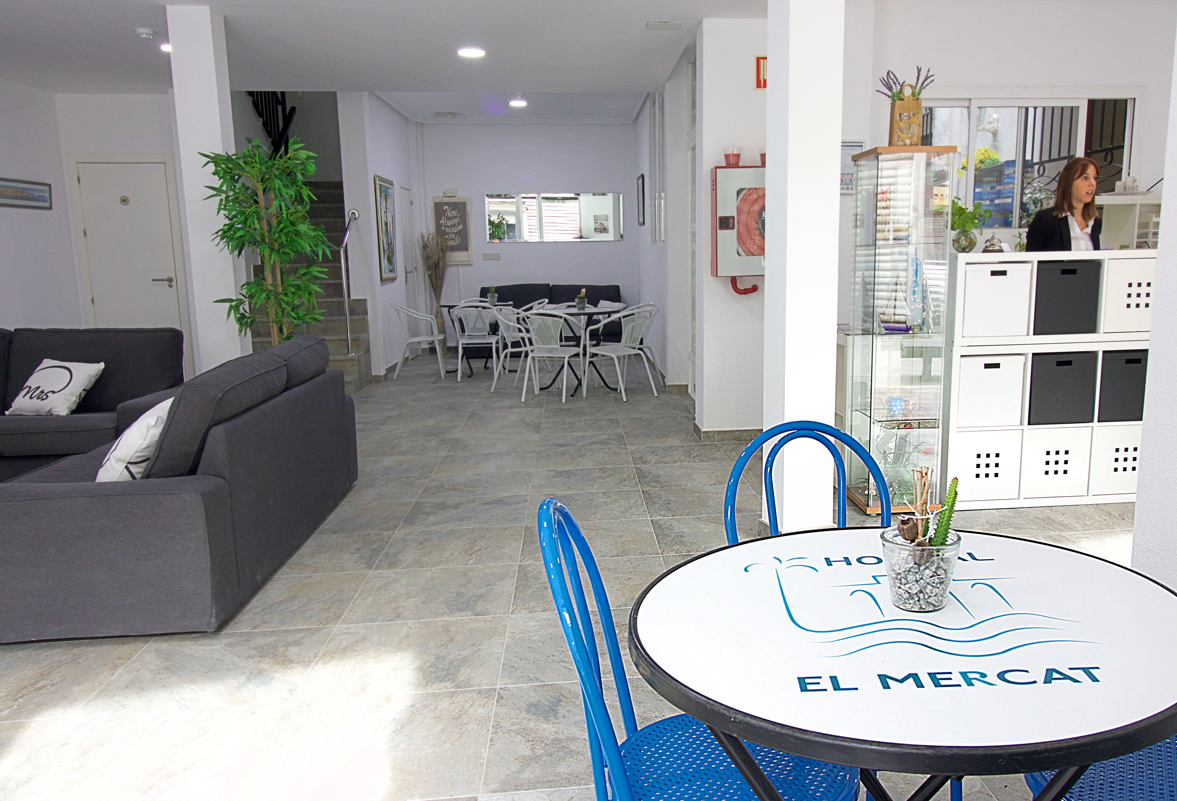 Una sala moderna con un sofá gris, un comedor con una mesa redonda con el letrero 'EL MERCAT', y una cocina con armarios y un refrigerador.