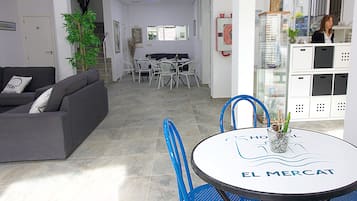 Una sala moderna con un sofá gris, un comedor con una mesa redonda con el letrero 'EL MERCAT', y una cocina con armarios y un refrigerador.