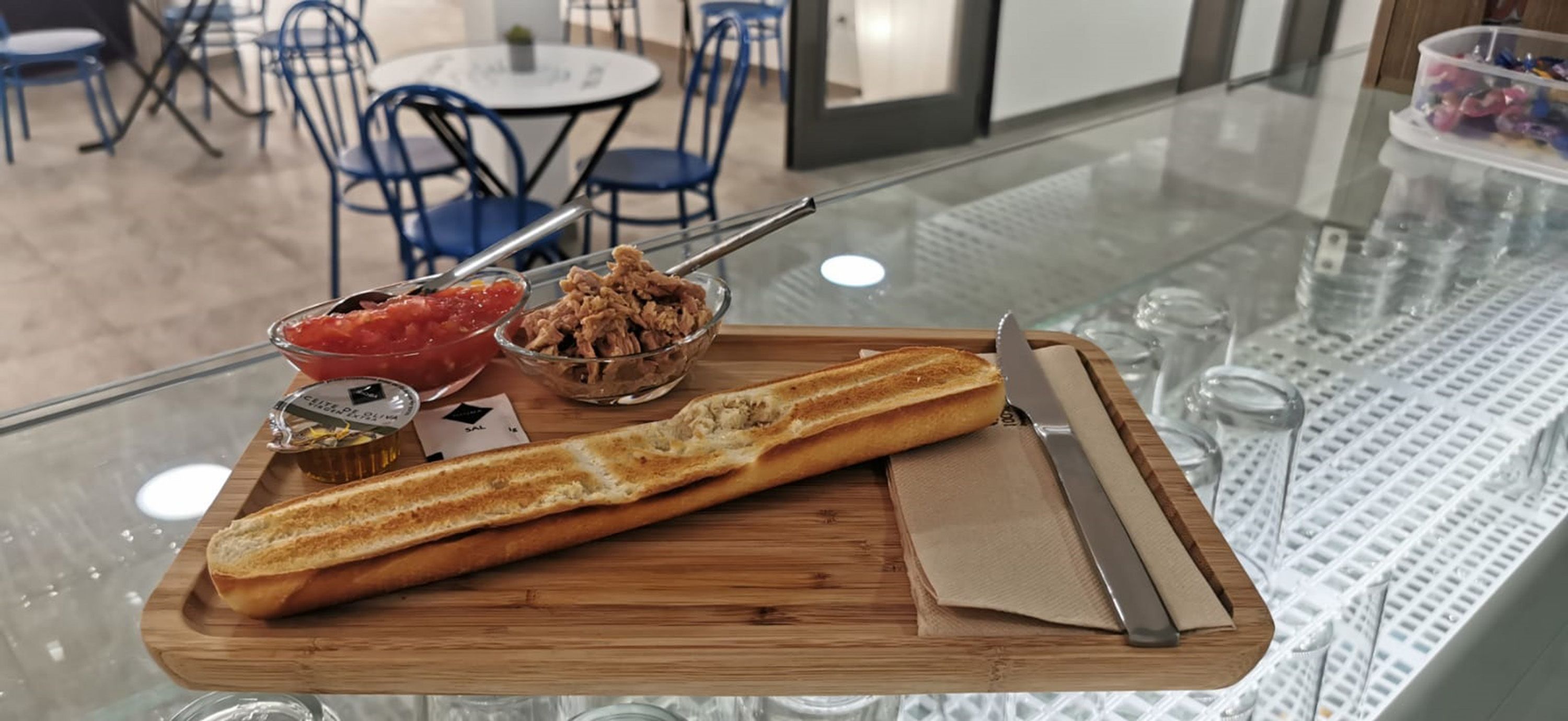 Ein Frühstücksbrett mit einem Baguette, einer Schüssel mit zerpflücktem Fleisch, einer Schüssel Tomatensalsa und einem Messer auf einem Glastisch.