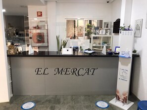 A reception desk with the name "EL MERCAT" prominently displayed. There are informational posters and a hand sanitizer dispenser on the counter. - Hostal El Mercat (Villajoyosa)