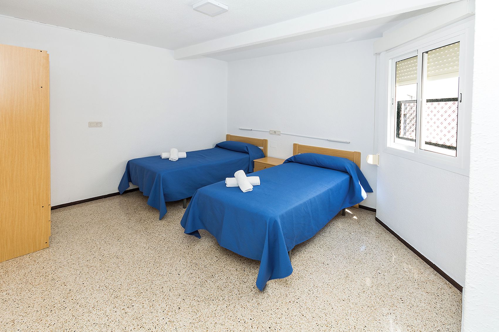 Una habitación con dos camas individuales, cada una con una colcha azul y toallas blancas, una puerta de madera y una ventana con persianas.