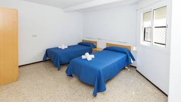 Una habitación con dos camas individuales, cada una con una colcha azul y toallas blancas, una puerta de madera y una ventana con persianas.