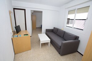 A small, sparsely furnished room with a wooden desk, a white table, and a grey sofa. There is a window with blinds, a door leading to another room, and a mirror on the wall. - Hostal El Mercat (Villajoyosa)
